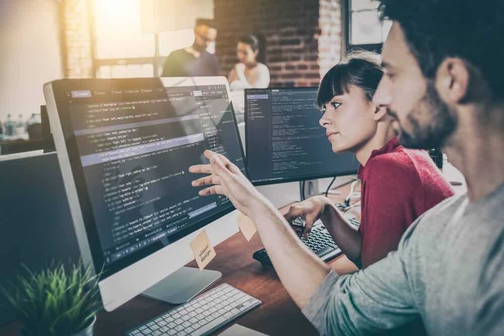 Two people collaborating on computer programming at a desk in a modern office setting with other colleagues in the background.