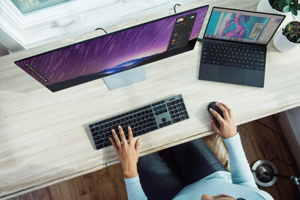 Person working at a desk with a large monitor, keyboard, and mouse, with a laptop next to the monitor displaying a colorful image. Plants and a window are in the background.