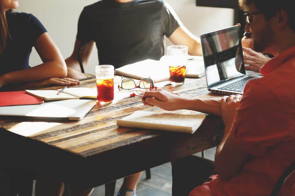 People sitting around a wooden table with notebooks, drinks, and a laptop in a casual meeting.