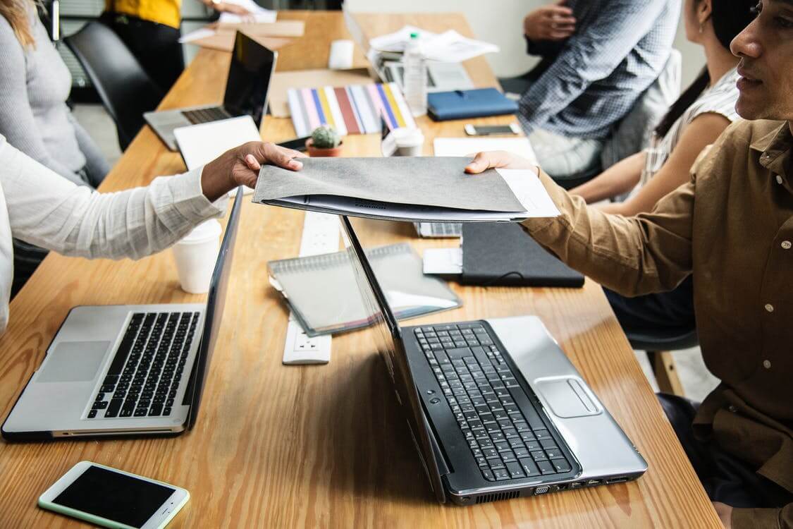 People exchanging a folder at a conference table with laptops, notebooks, and papers around them.