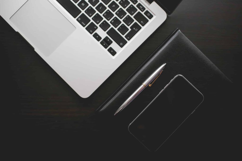 Laptop, pen, notebook, and smartphone on a dark wooden desk.