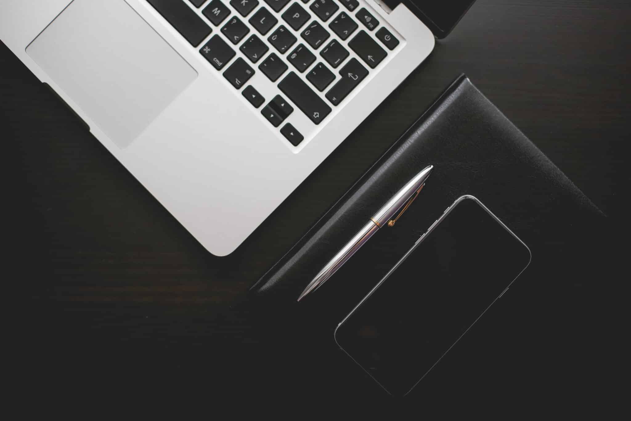 Laptop, pen, notebook, and smartphone on a dark wooden desk.