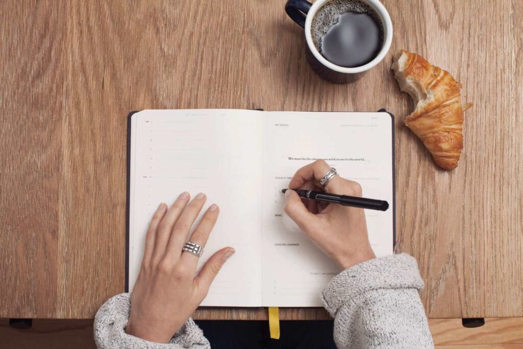 A person writes in a notebook with a pen on a wooden table. A cup of black coffee and a croissant are nearby. The person is wearing rings and a cozy, light-colored sweater.