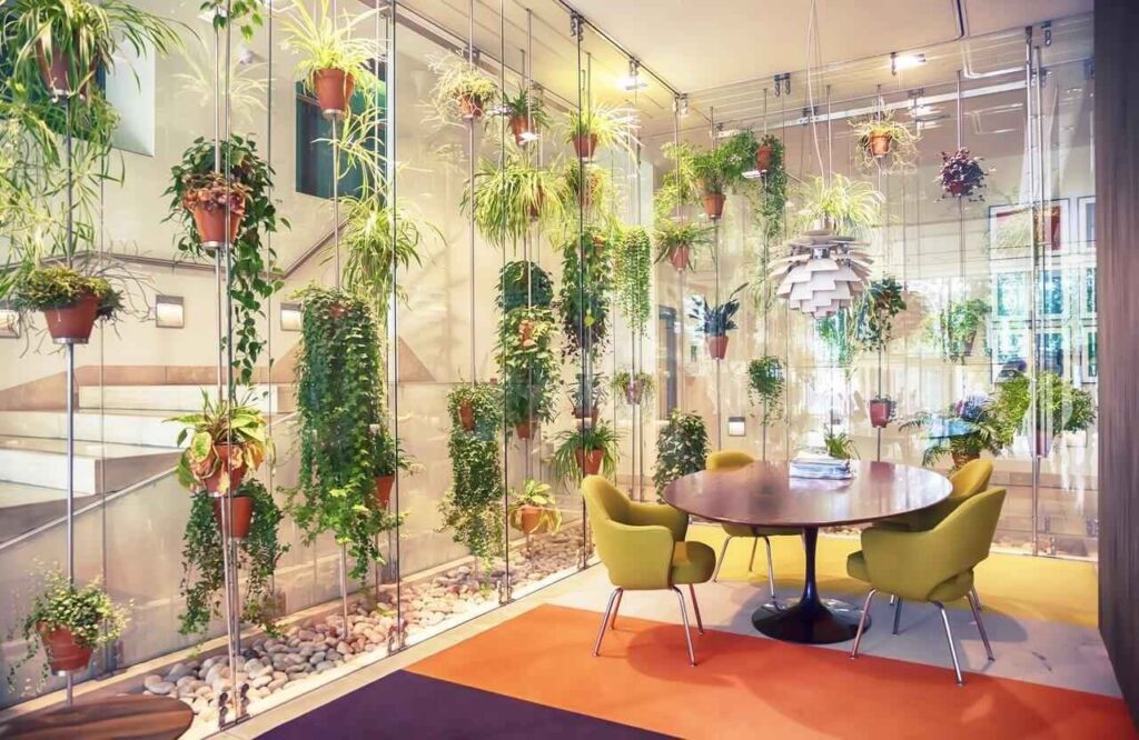 Modern room with hanging plants, a round table, and green chairs on a colorful carpet. Glass walls surround the space.
