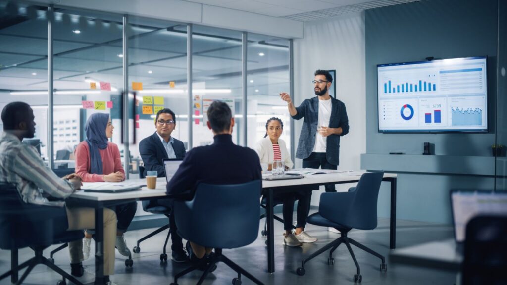 A man presents data on a screen to colleagues seated around a conference table in a modern office.