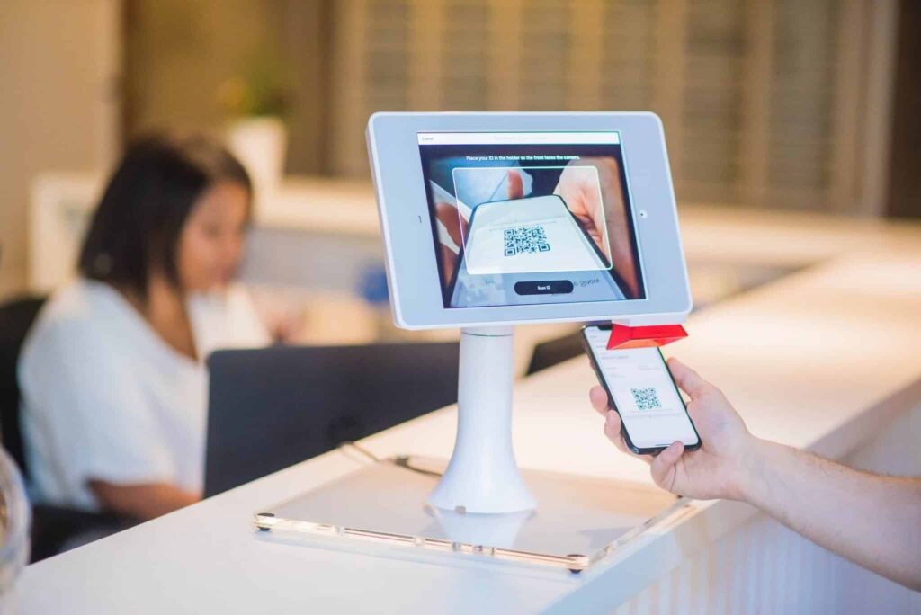 A person scans a QR code on a phone at a kiosk; a woman works on a laptop in the blurred background.