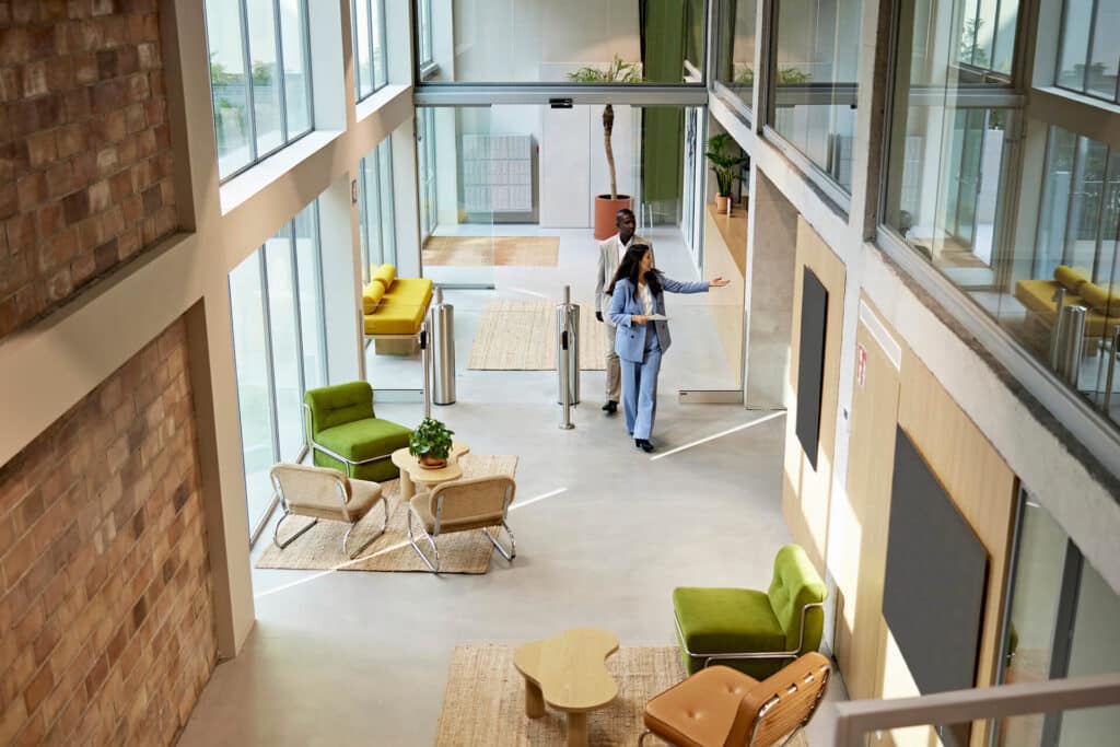 Modern office lobby with glass walls, green chairs, and two people walking through the space.