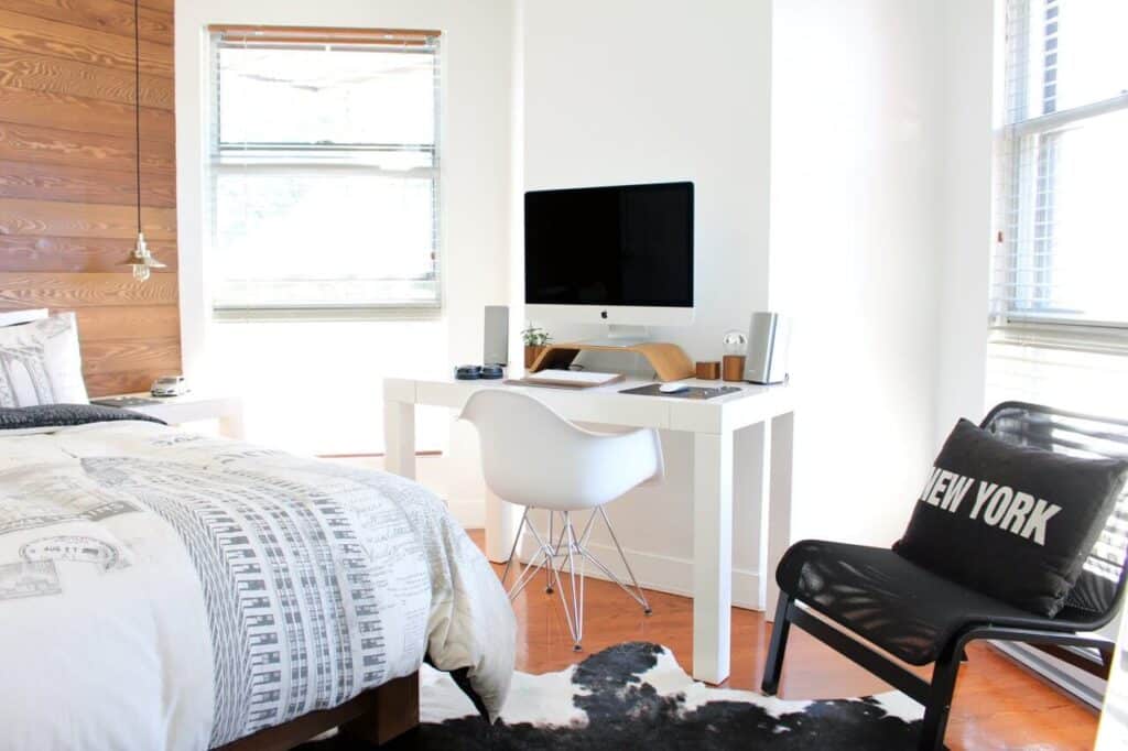 Bright bedroom with a desk, computer, chair, and "New York" pillow on a black chair, next to a bed.