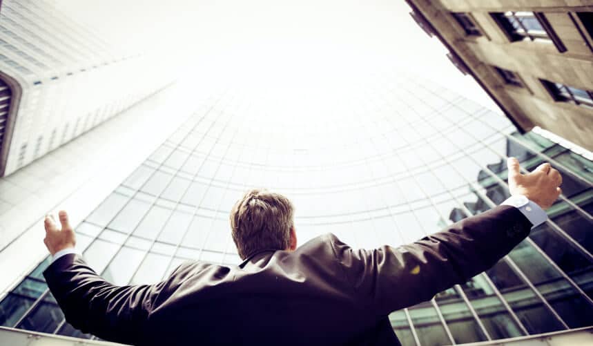 Man in a suit standing with arms outstretched, gazing up at tall modern buildings.