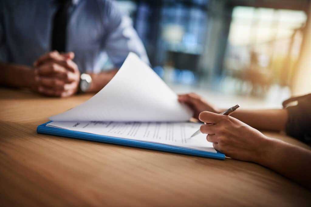Two people sitting at a table, one holding a pen over documents with a blue folder in an office setting.