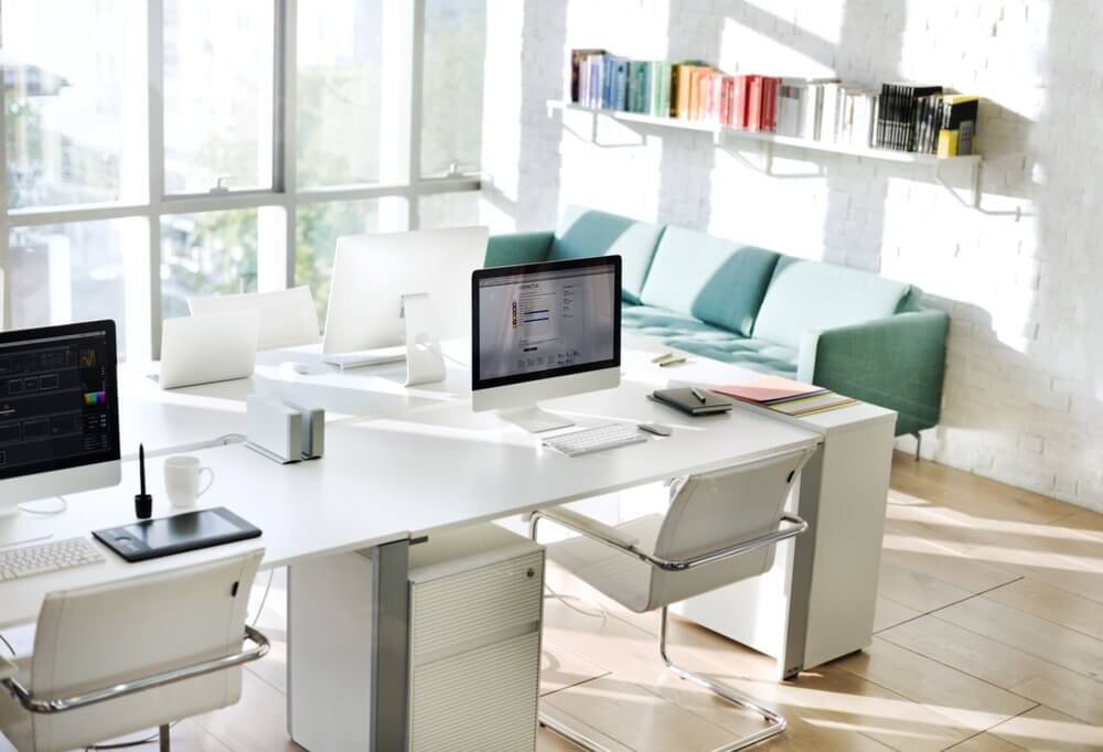 Modern office with white desks, computers, chairs, large windows, green couch, and wall shelves with colorful books.