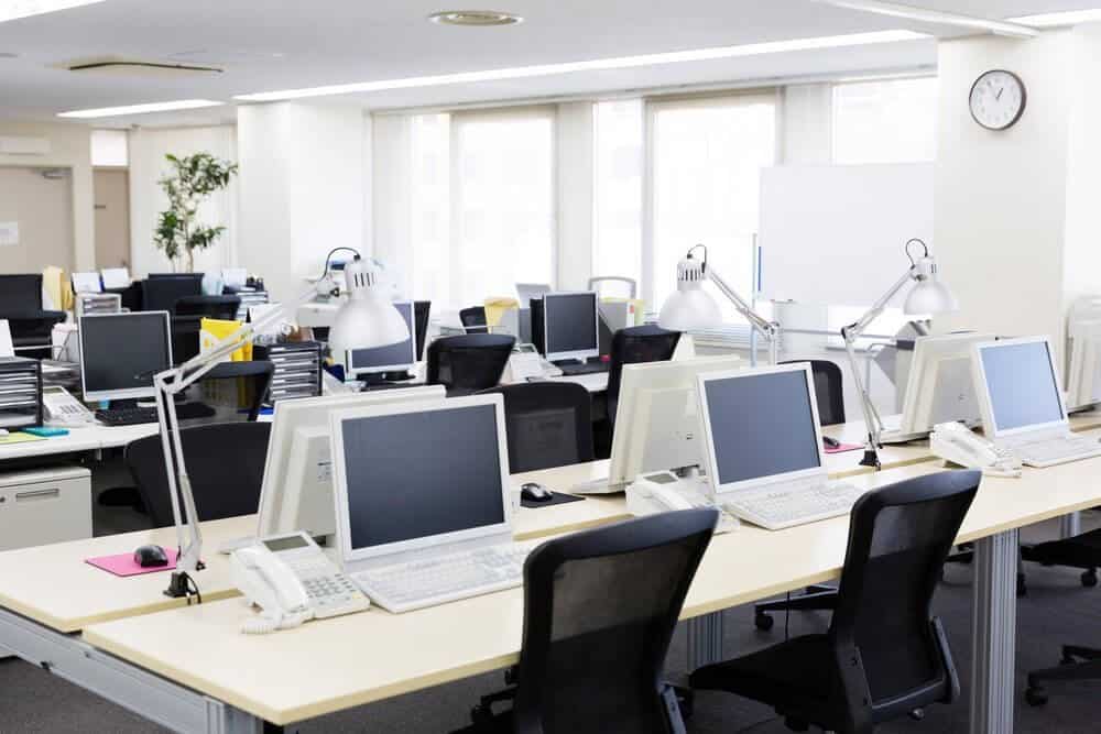An empty, modern office with rows of desks, computers, lamps, and chairs under bright fluorescent lighting.