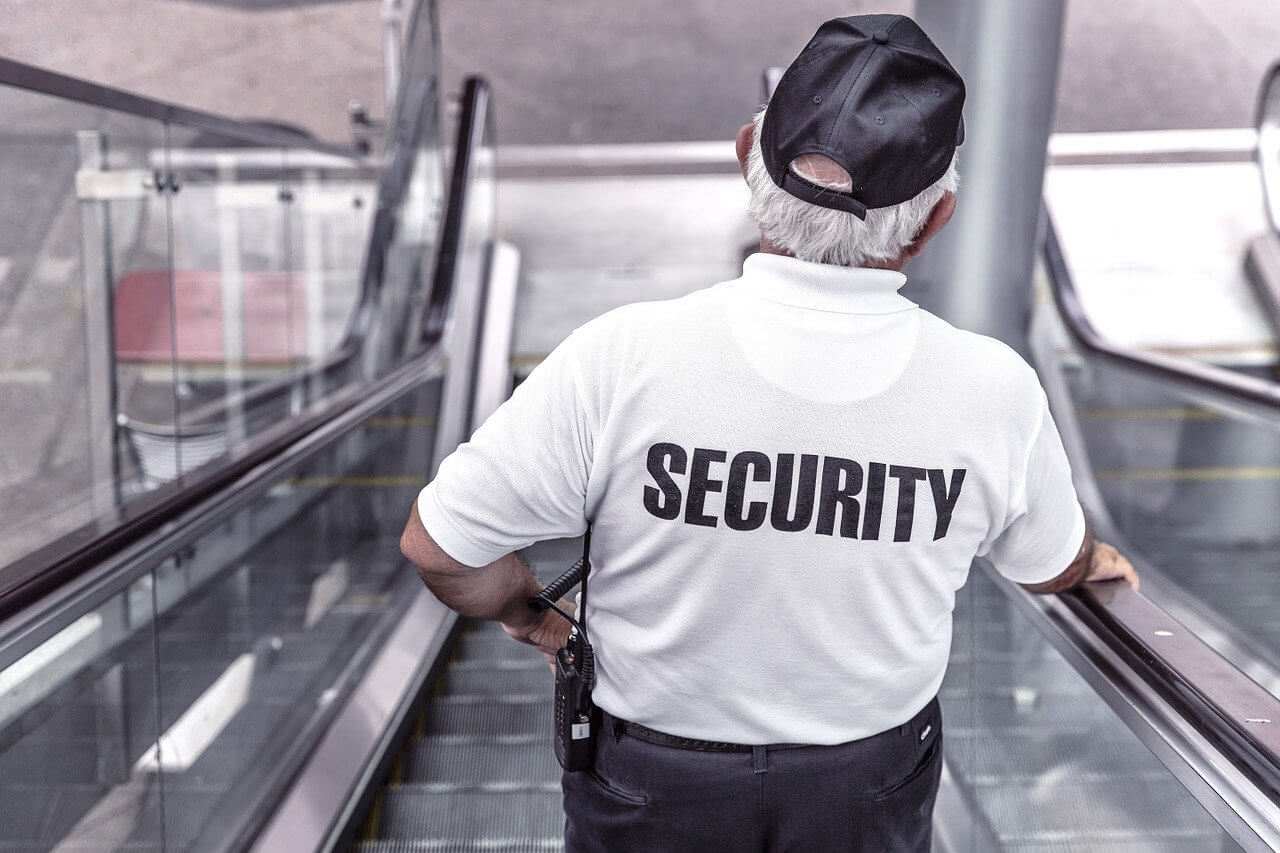 Security guard wearing a white shirt and black cap, standing on an escalator, looking downwards.