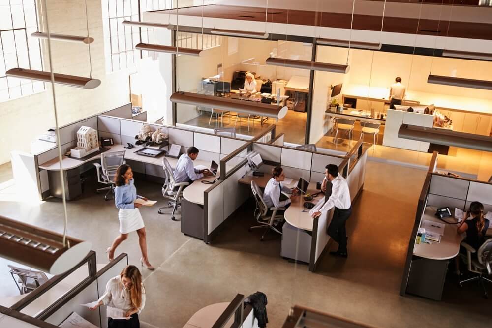 Modern office with people working at desks in cubicles, large windows, and overhead lighting.