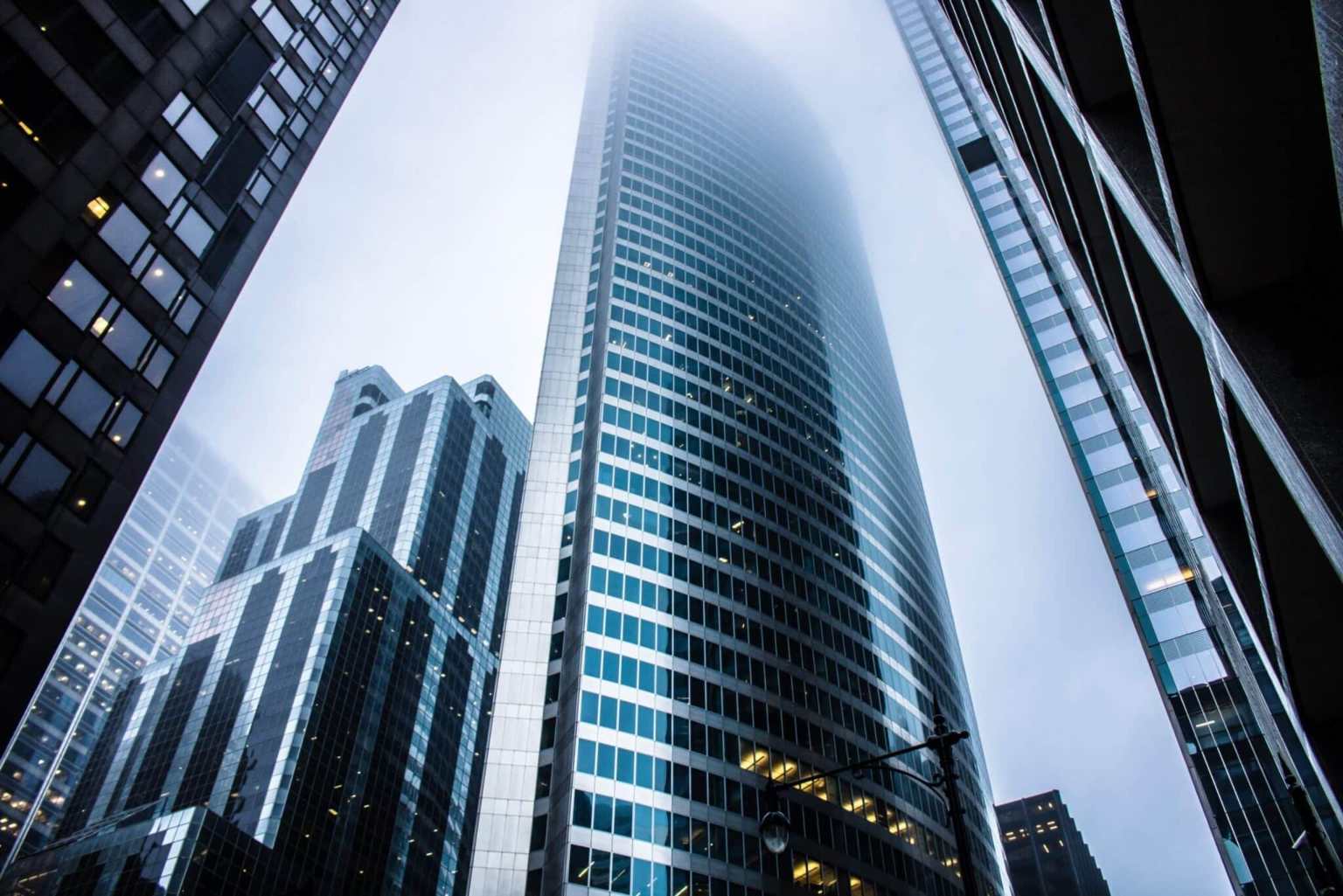 Tall modern skyscrapers with glass windows rise into a foggy sky, with one building centrally framed and partially obscured by mist. Other high-rises outfitted with advanced office sensors surround it, and a streetlight is visible at the bottom.