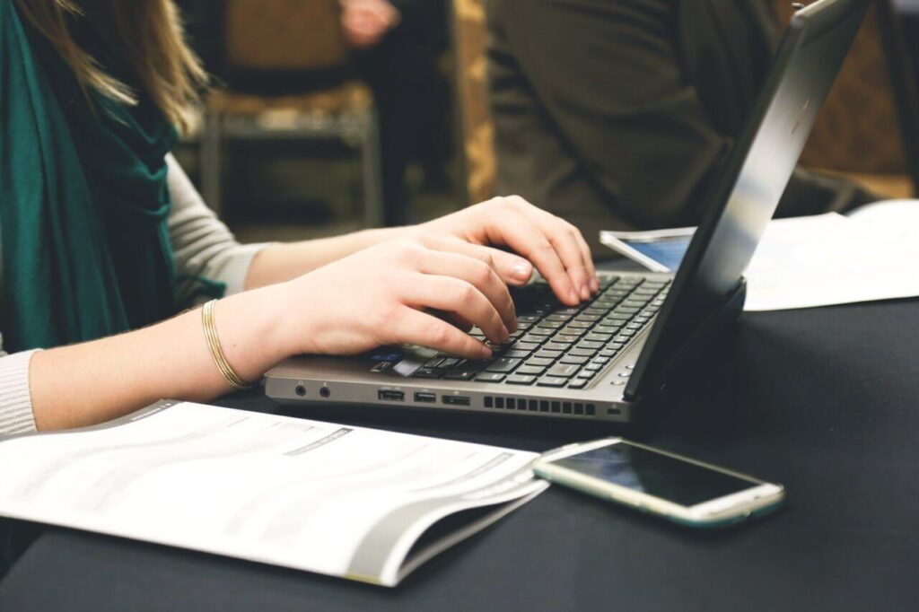 Person typing on a laptop with documents and a smartphone nearby on a table.