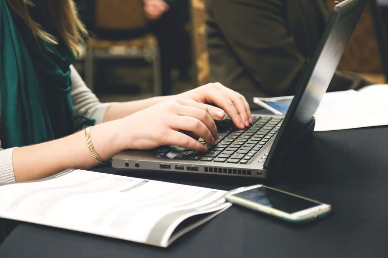 Person typing on a laptop with documents and a smartphone nearby on a table.