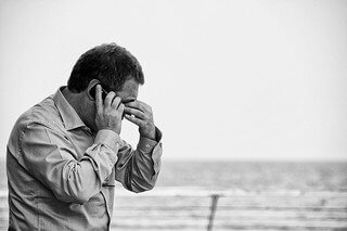 Person on a phone call near the ocean appears stressed, holding their forehead with their hand.