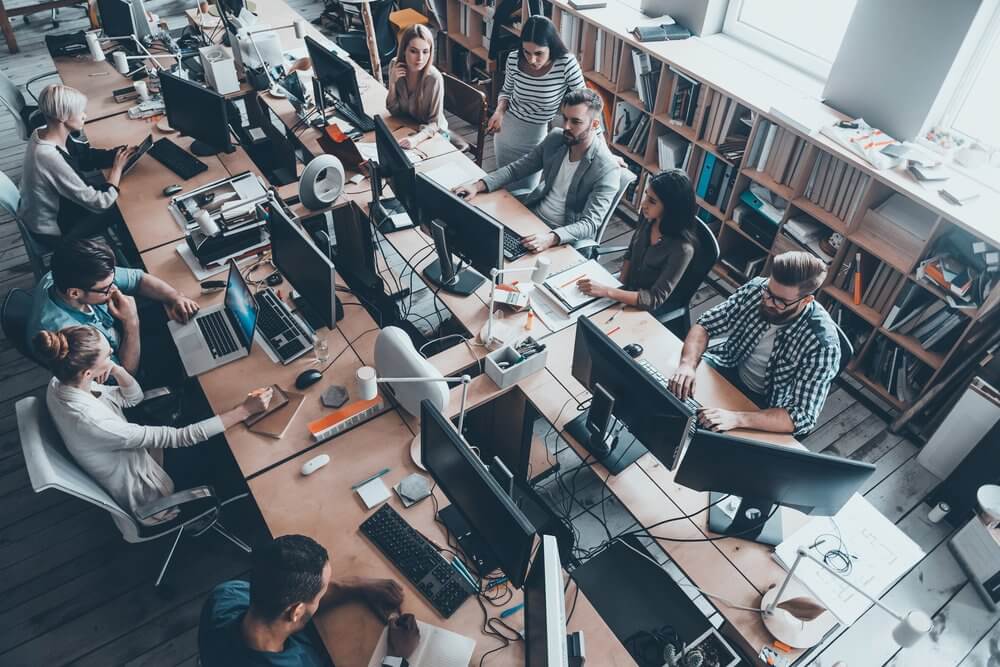 Aerial view of a busy office with colleagues working at long desks with computers and bookshelves in the background.