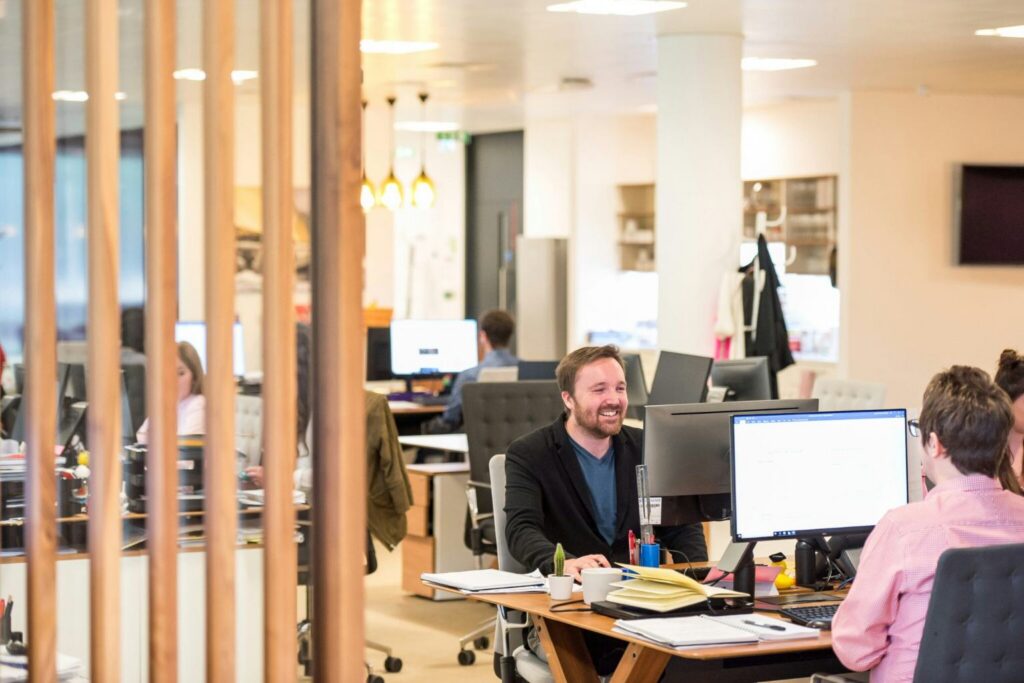 Open-plan office with people working at desks. Two men are smiling and talking across computer monitors, while others in the background focus on their tasks. The workspace features modern furniture, warm lighting, and thoughtful office design that encourages collaboration and productivity.