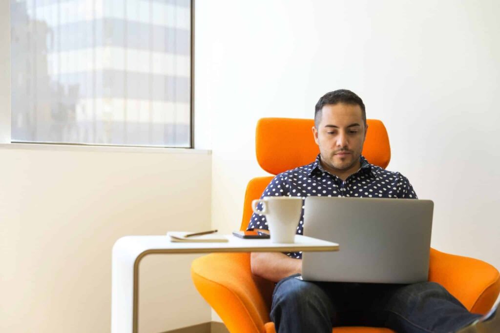 A man sits in an orange chair working on a laptop, embracing activity-based working as he chooses a comfortable spot to focus. A white table beside him holds a smartphone, notebook, and mug, while a large window in the background lets in natural light.