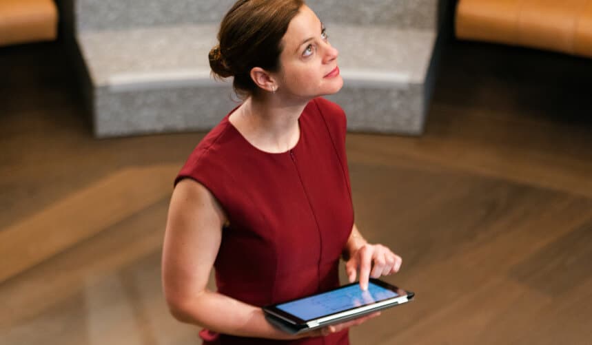 Woman in a red dress looks upward while holding a tablet, standing in a modern indoor space.