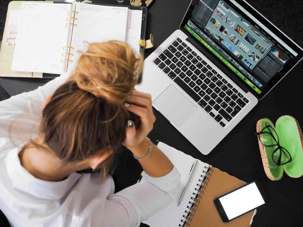 A person sits at a desk with their head in their hands, surrounded by a laptop, notebooks, a phone, glasses, and papers, appearing stressed or overwhelmed by work.