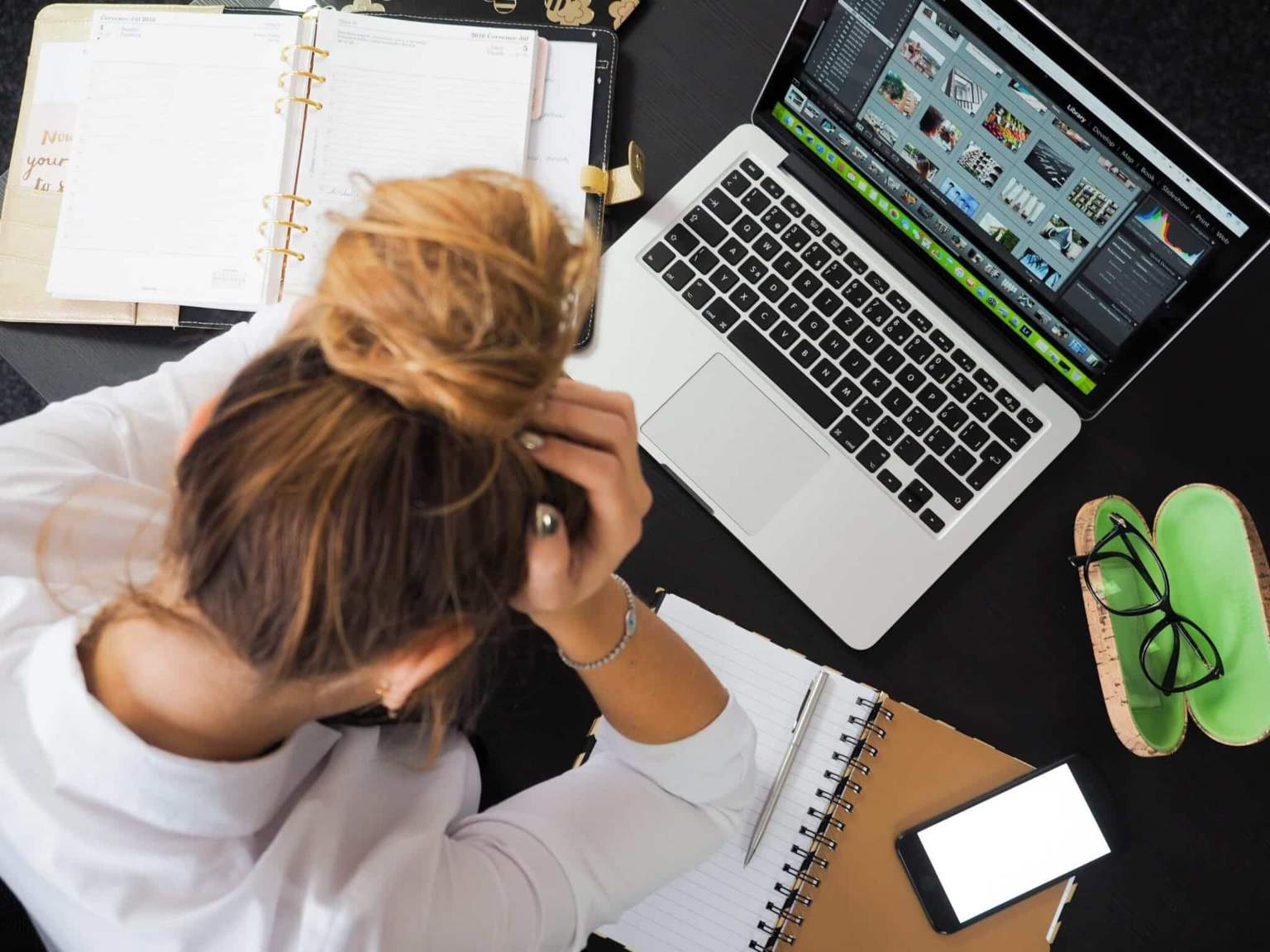 A woman with her hair in a bun sits at a desk, holding her head in her hands, surrounded by a laptop, open notebooks, a smartphone, glasses case, and papers.
