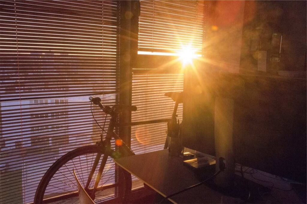 Sunlight streams through window blinds, illuminating an indoor scene with a bicycle and desk, creating a warm, glowing atmosphere with visible sun rays and shadows across the room.