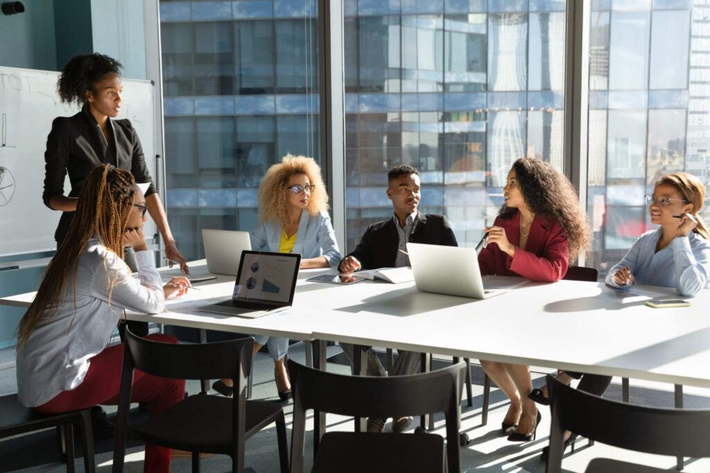 Six people sit around a conference table with laptops and papers, having a discussion in a modern office with large windows. One person stands by a whiteboard presenting room booking analytics, while the others listen and take notes.