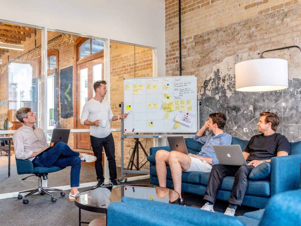 Four members of the ifm team gather in a modern office to discuss ideas. One stands by a whiteboard covered in sticky notes, while three sit on a blue sofa with laptops, listening attentively. Exposed brick walls and large windows are visible in the background.