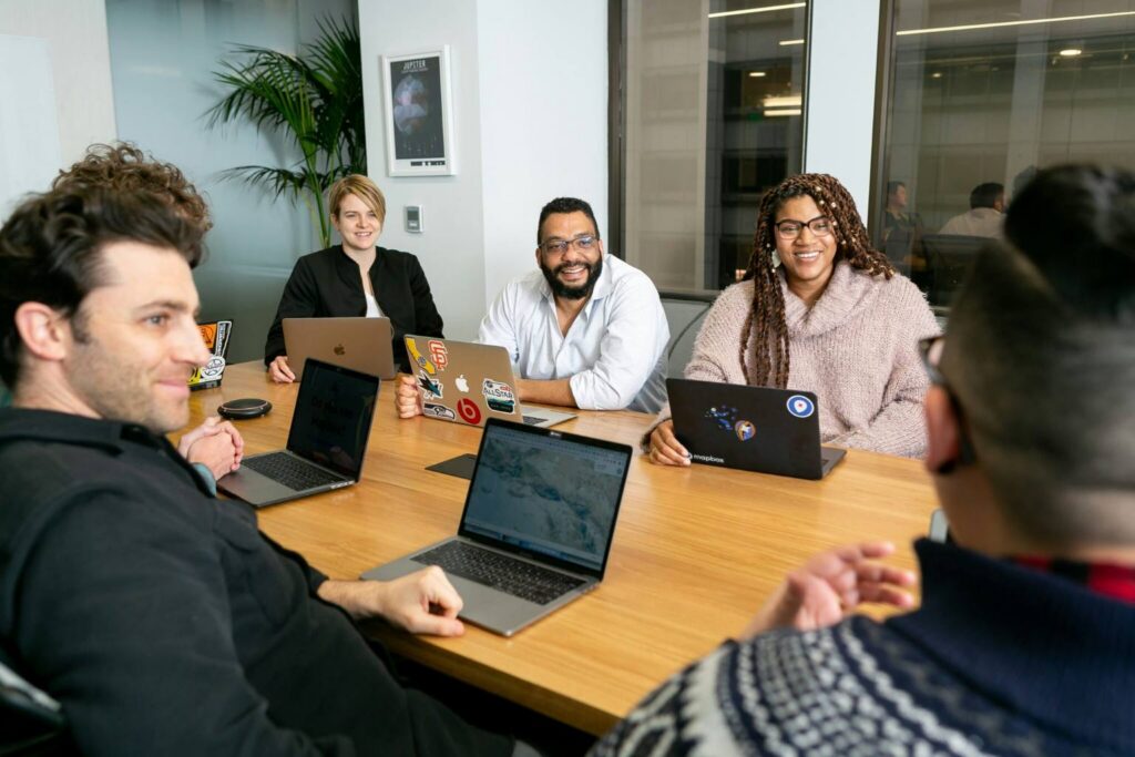 Five people smiling around a conference table with laptops in a modern office setting.