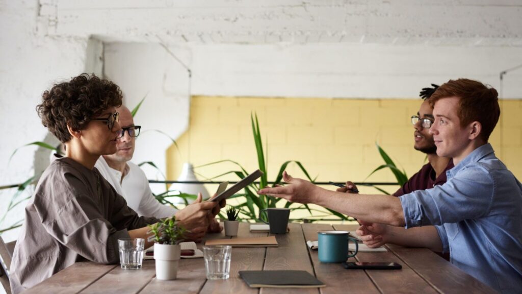 Four people from different generations in the workplace sit at a wooden table with plants, mugs, and papers, having a meeting. One person is handing a tablet to another while others watch and listen.