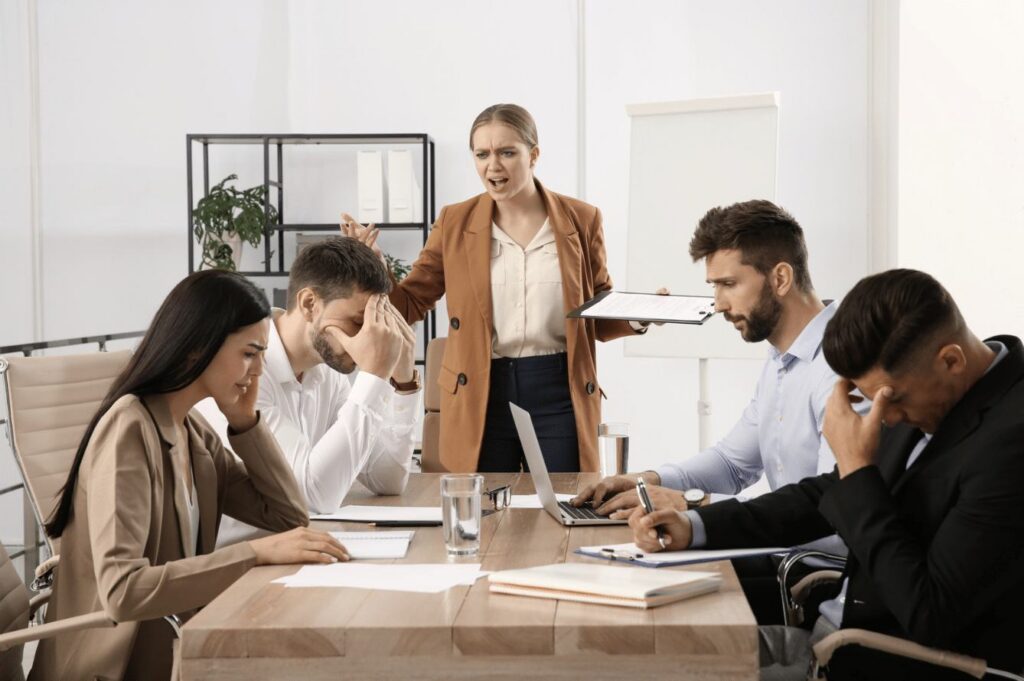A woman stands and gestures at a stressed group of four people sitting at a conference table.