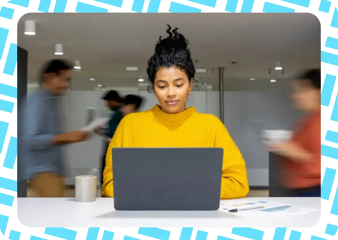 A person in a yellow sweater works on a laptop at a desk with papers and a mug, while blurred figures move in the background. The image has a blue geometric border.