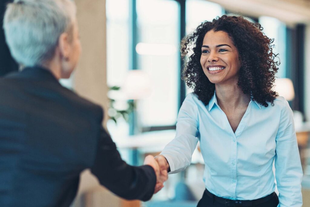 Two women shaking hands in a bright office, one smiling in a light blue shirt, the other with short gray hair