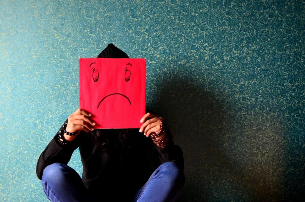 Person sitting against a teal wall, holding a red paper with a drawn sad face covering their own face.