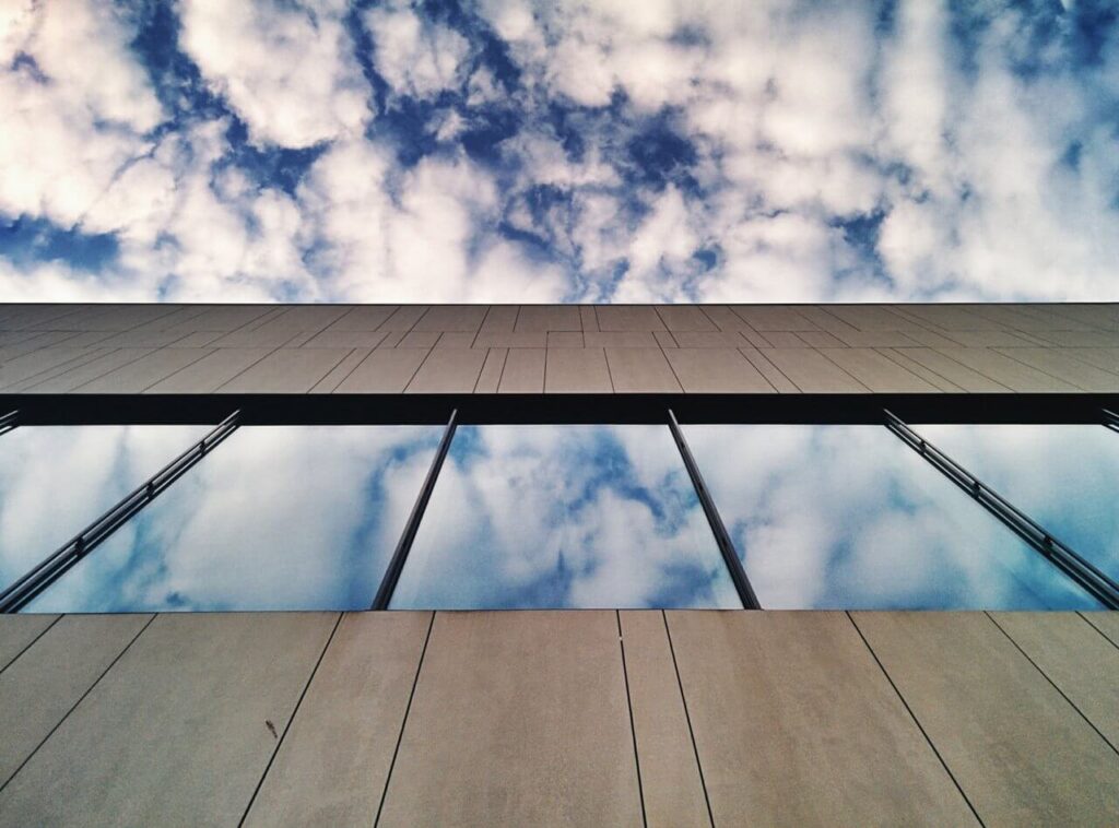Modern building facade with large glass windows reflecting a partly cloudy sky, viewed from below. The sky above the building is blue with scattered white clouds.