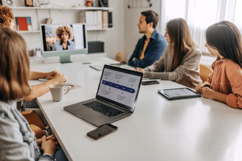 Five people have a meeting in a bright office, reflecting the return to office trend; four sit at a table with laptops and tablets, while one joins via video call. A flowchart is displayed on a laptop in the foreground.