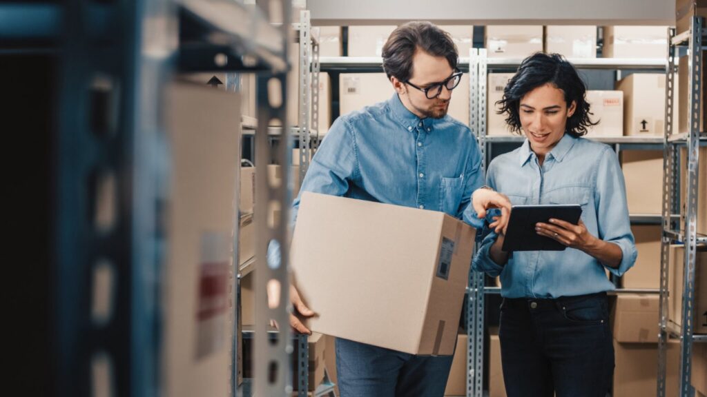 Two people in a warehouse, one holding a box, the other pointing at a tablet, standing among shelves of cardboard boxes.