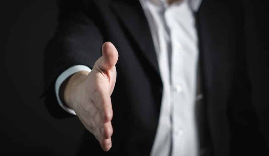 Close-up of a person in a suit extending a hand for a handshake, against a dark background.