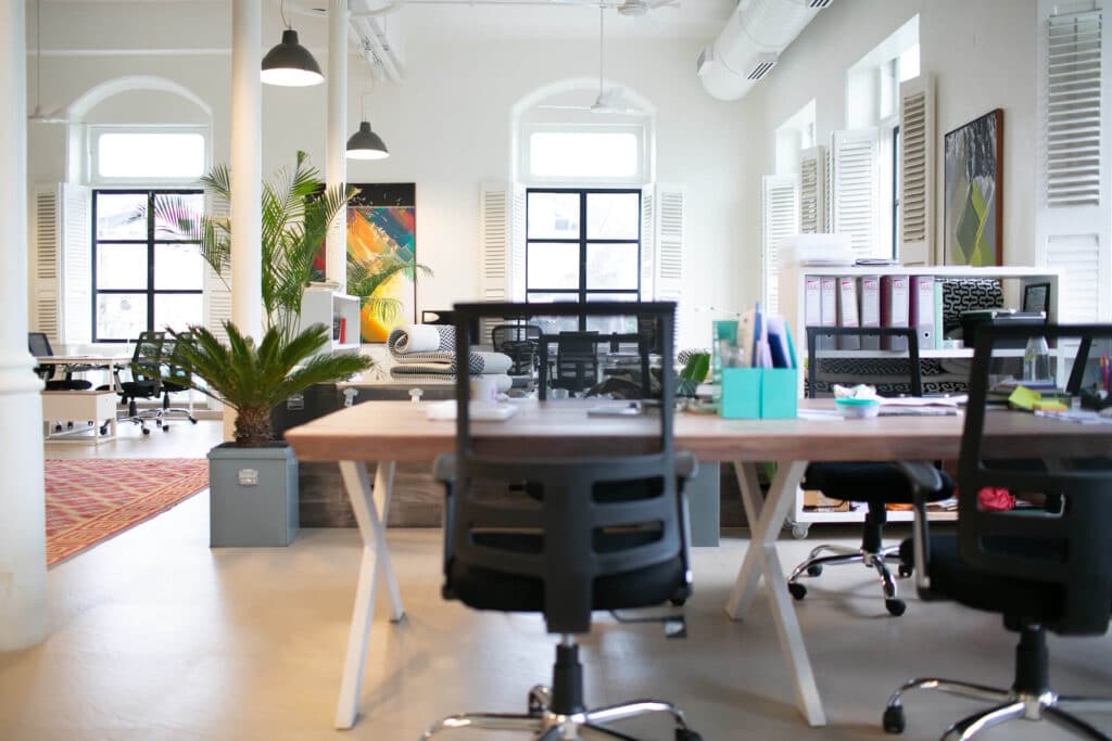 Open-plan office with modern furniture, plants, and colorful wall art, featuring large windows and natural light.