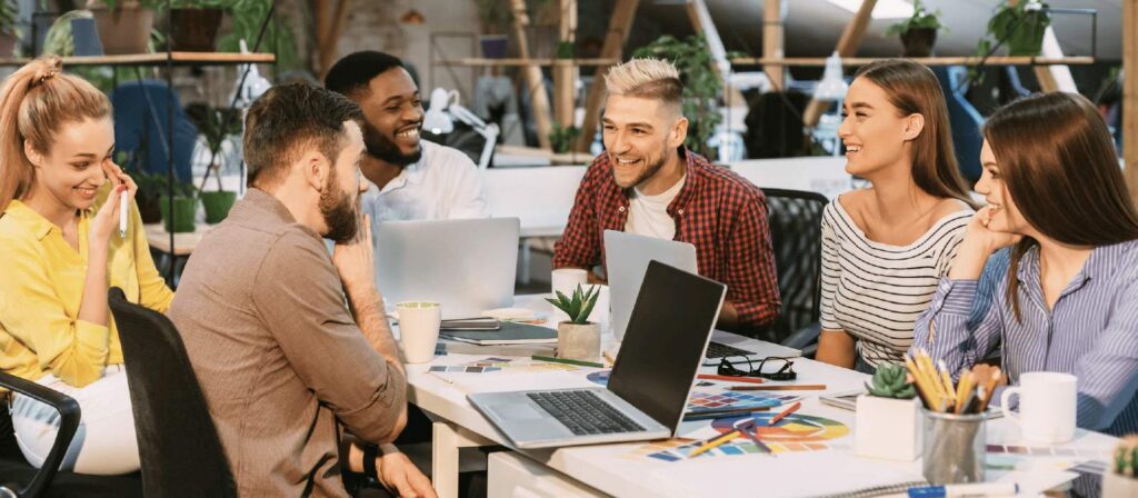 Group of young professionals in casual attire gather around a laptop in a modern workplace