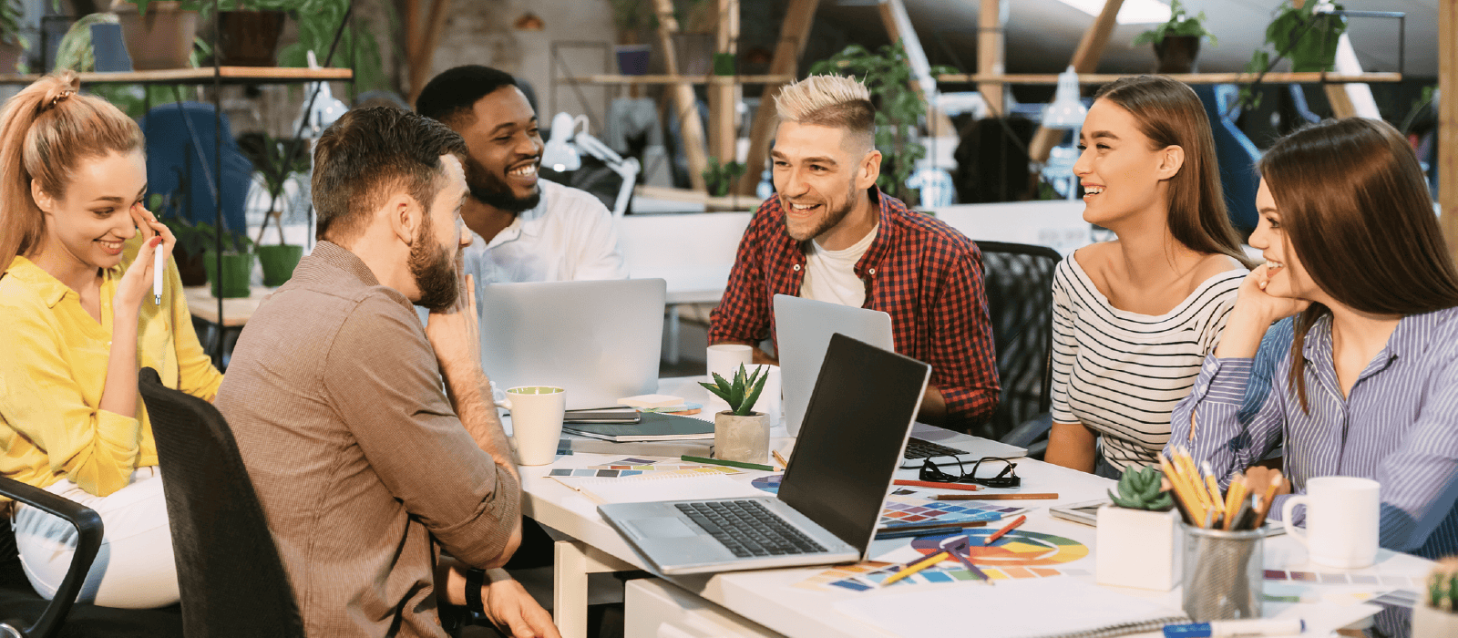 Group of young professionals in casual attire gather around a laptop in a modern workplace
