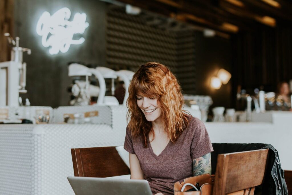A woman with wavy red hair, wearing a purple shirt, smiles while using a laptop in a modern coffee shop with a bright sign and barista equipment in the background, embodying the dynamic energy of multiple generations in the workplace.