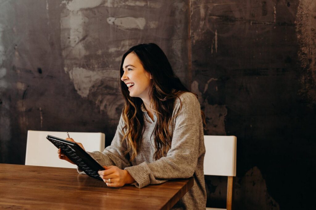 A woman with long hair, wearing a gray sweater, sits at a wooden table holding a tablet and smiling, clearly enjoying the benefits of new technology in the workplace, with a dark, textured wall in the background.
