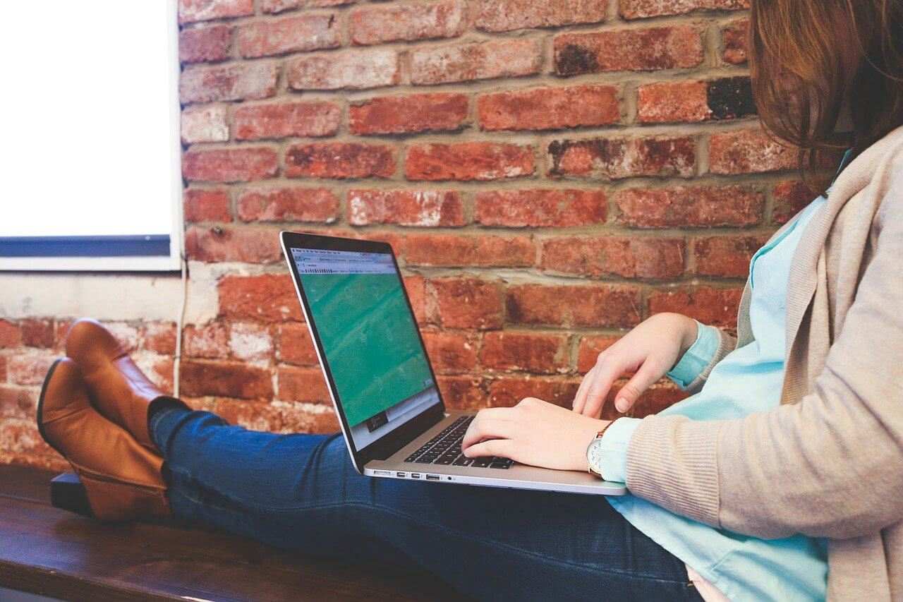 Person sitting on a bench, typing on a laptop, with a brick wall in the background.