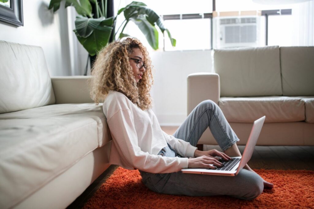 A person with curly hair and glasses sits on an orange rug, leaning against a white couch, typing on a laptop in a bright living room with plants and large windows—perhaps researching or reflecting on the dynamics of generations in the workplace.