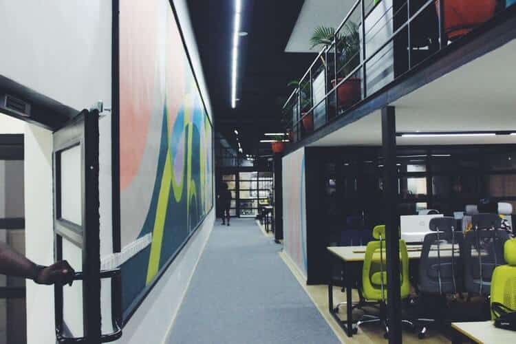 Modern office hallway with open-plan workstations and green chairs on one side, abstract art on the opposite wall.