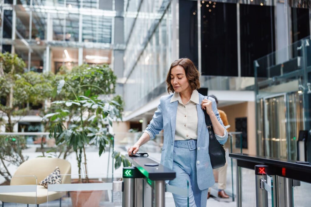 A businesswoman in smart casual attire confidently walks through security turnstiles into a glass-structured office building, ready for the workday.